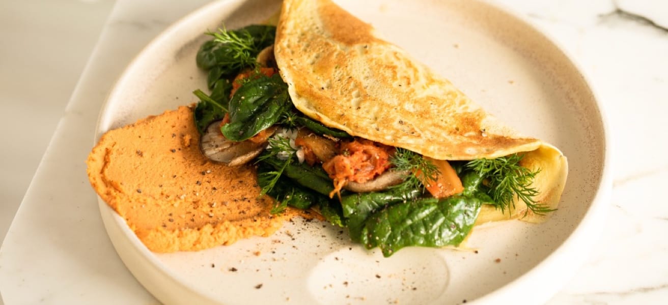 A folded crepe with on a plate, filled with vegetable matter. Also on the plate is some orange humus